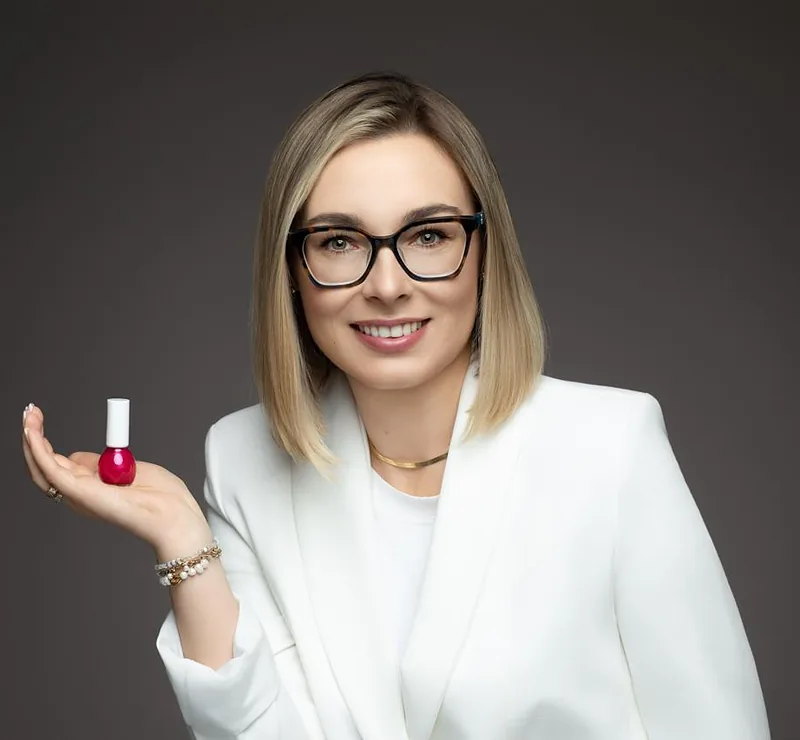 Justyna Krysiak w białej marynarce, na dłoni trzyma buteleczkę czerwonego lakieru